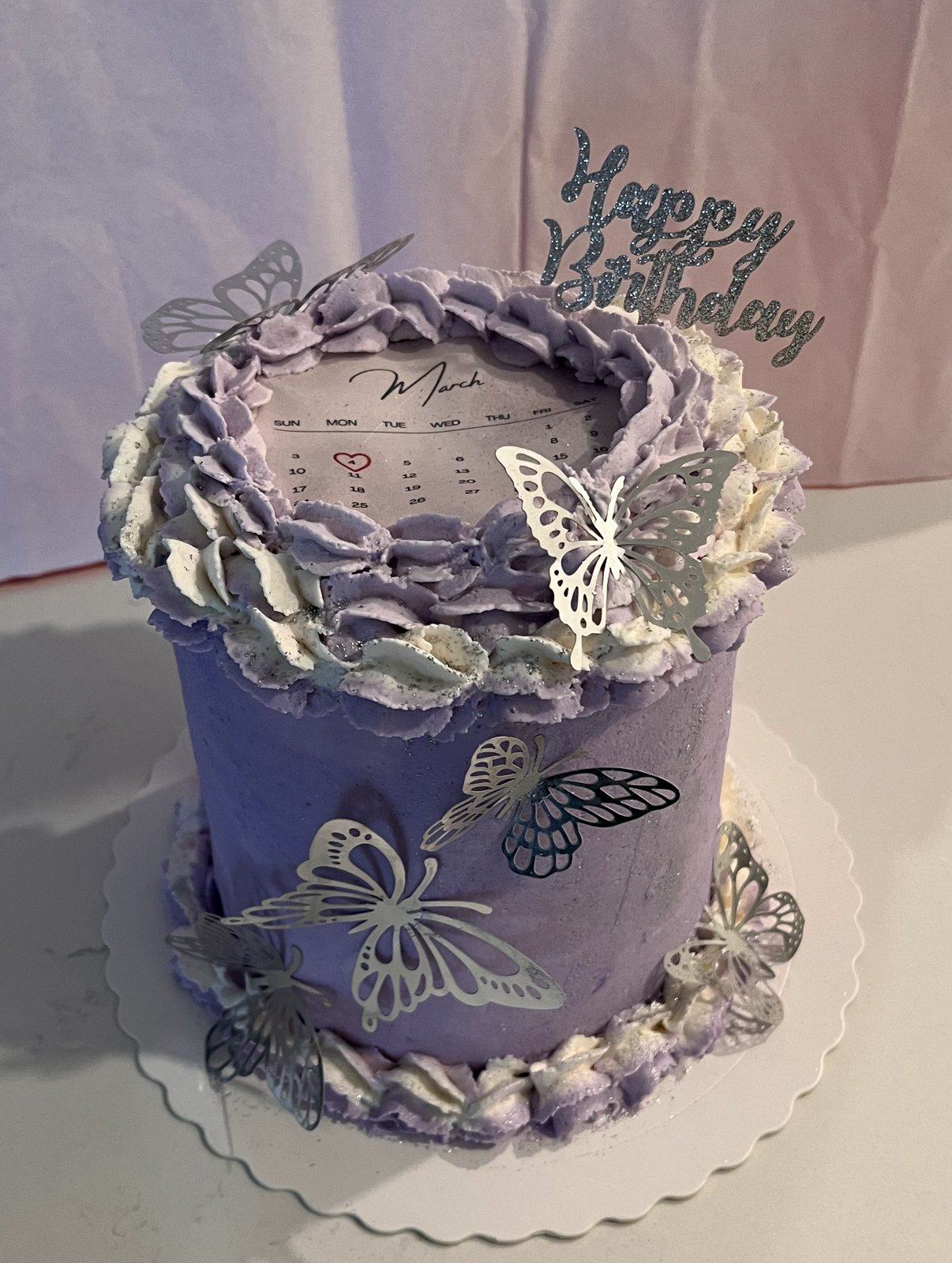 Tall lavender buttercream cake with a printed calendar on top marking the birthday, piped ruffles, silver butterflies, and a glitter Happy Birthday topper.