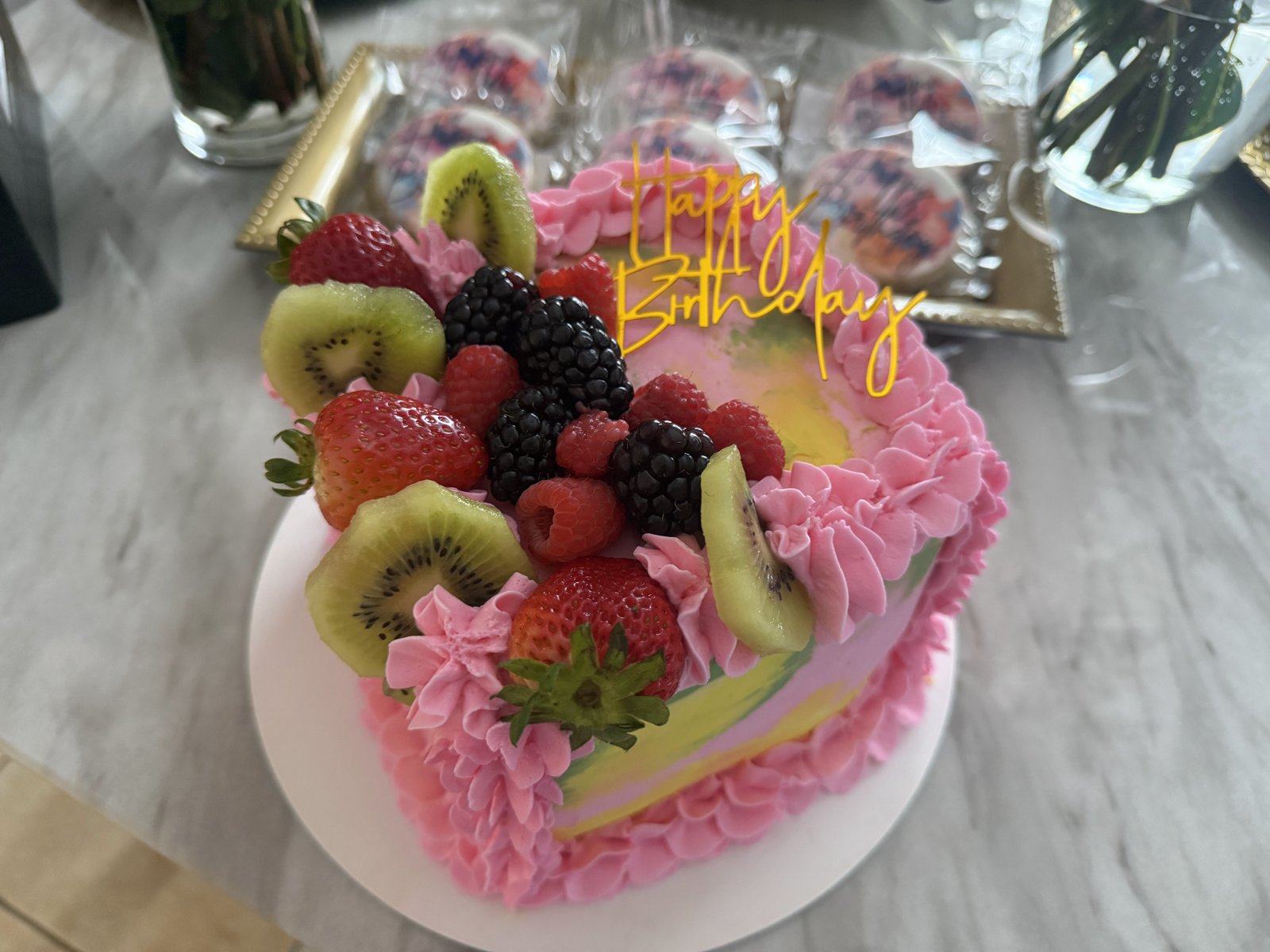 Pink buttercream birthday cake topped with kiwi, strawberries, and blackberries, with a gold Happy Birthday topper.