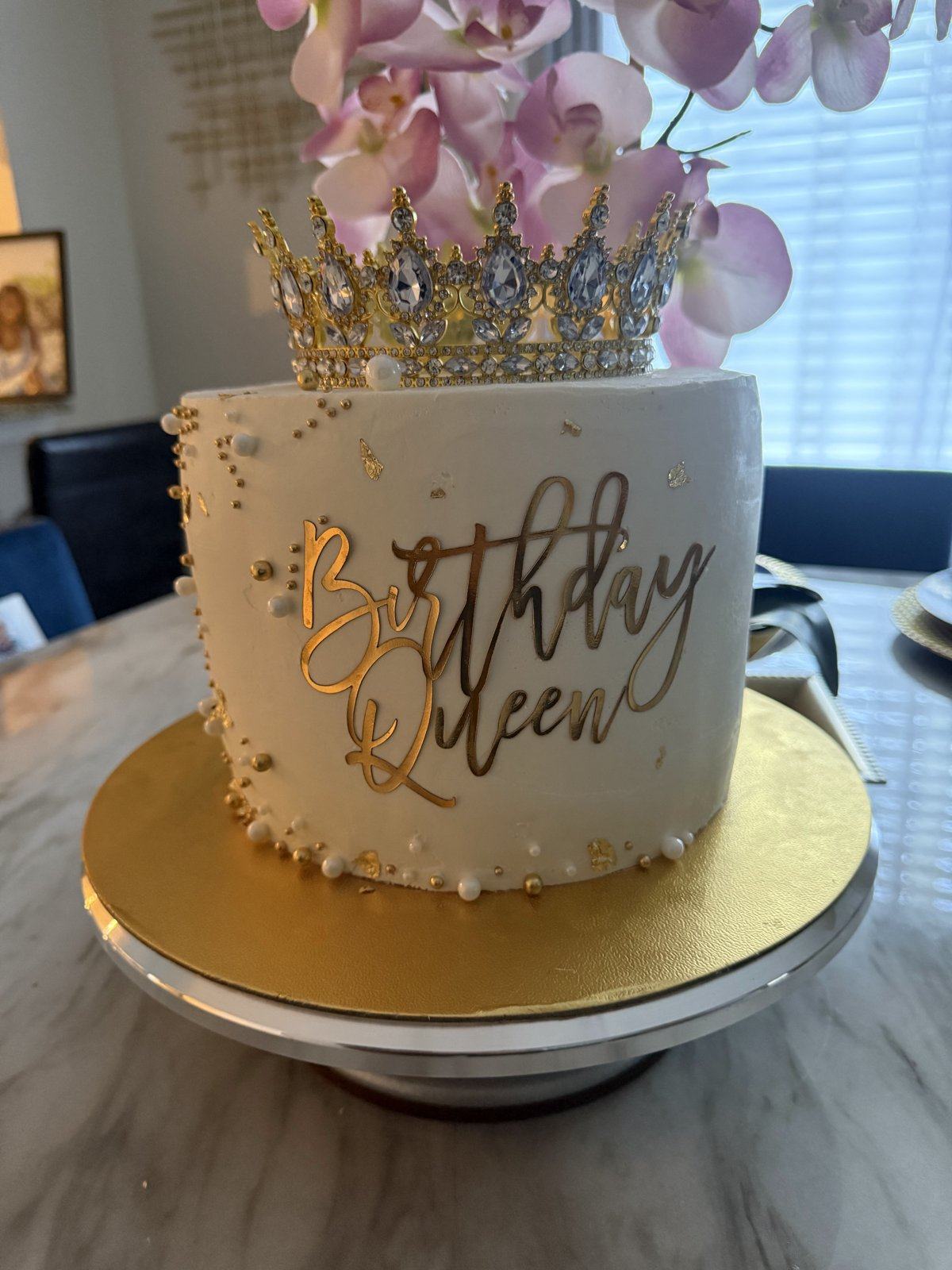 White birthday cake with gold Birthday Queen lettering, gold pearls, and a jeweled crown on top.