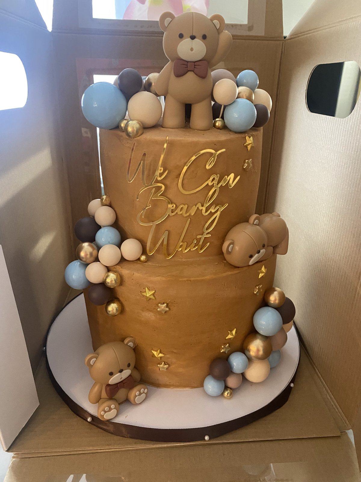 Two-tier caramel baby shower cake reading We Can Bearly Wait, with fondant teddy bears and blue, brown, and gold spheres.