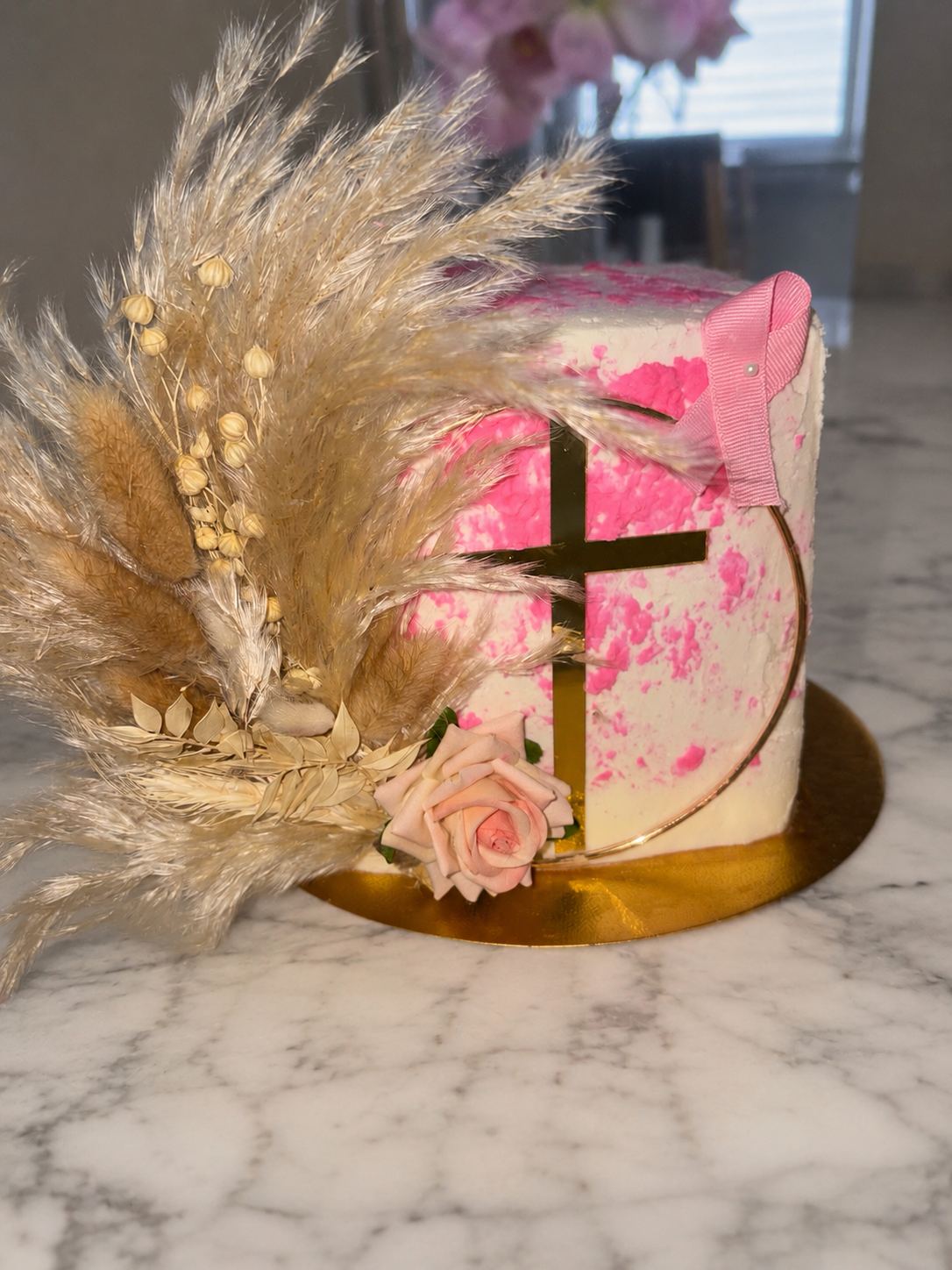 White cake with pink texture, a gold cross inside a gold hoop, a pink bow, a soft pink rose, and a spray of dried pampas grass.