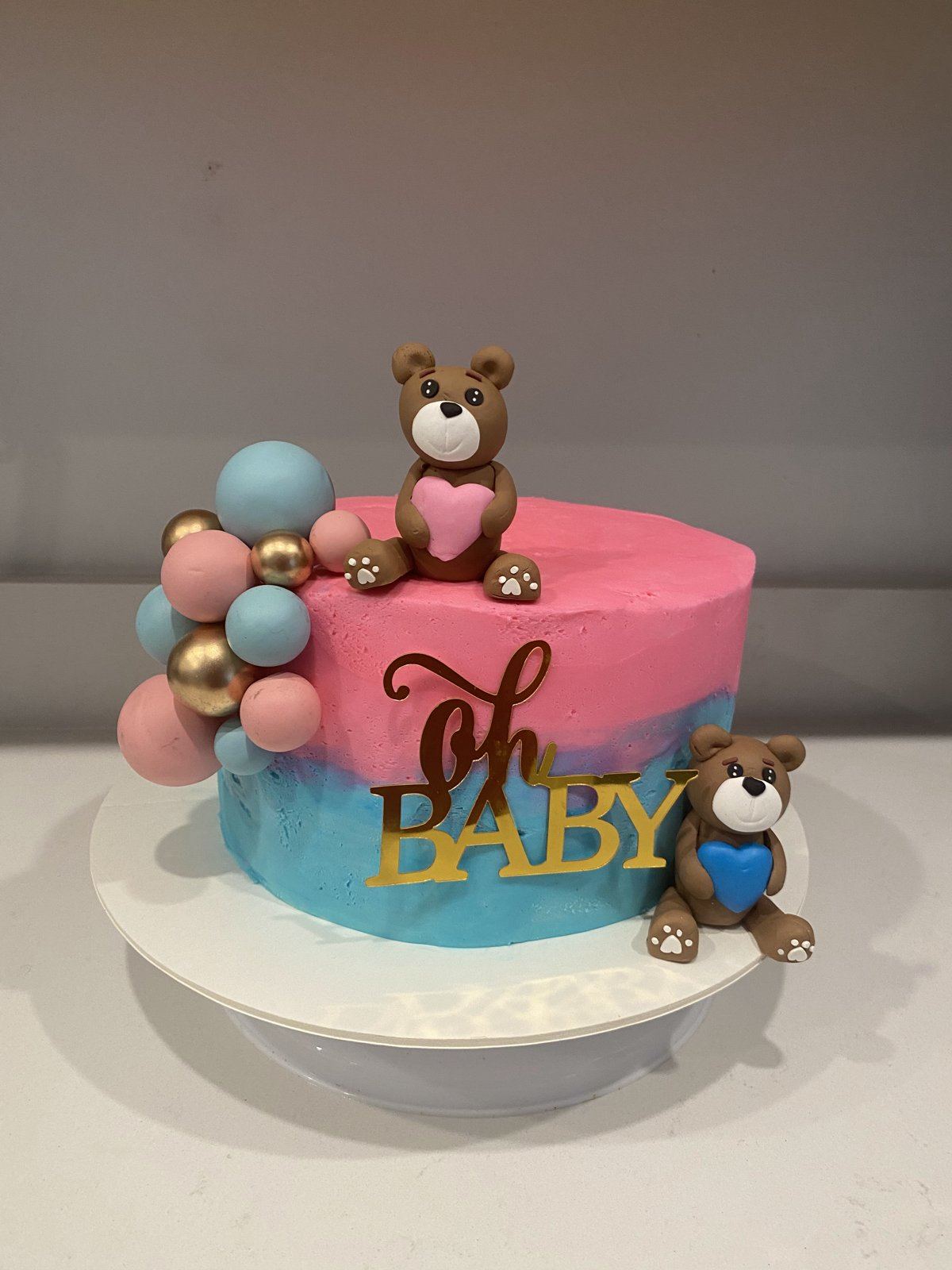 Gender reveal cake frosted half pink and half blue with gold Oh Baby lettering, pink and blue balloon spheres, and two fondant teddy bears.