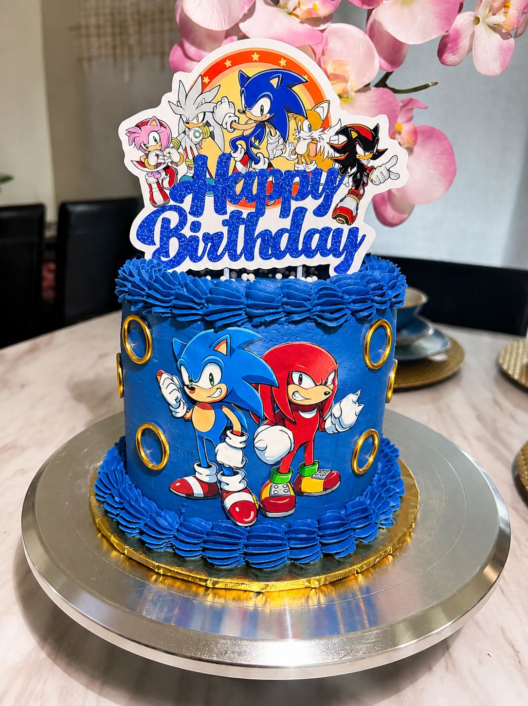 Blue buttercream birthday cake with gold ring decorations, cartoon hedgehog character images, and a glittery Happy Birthday topper.