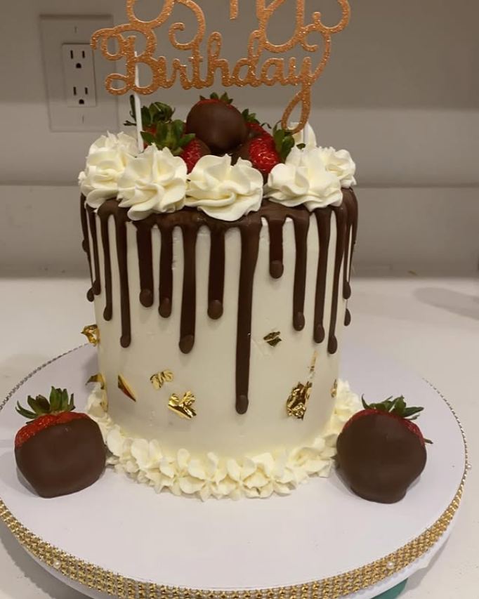 Tall white cake with chocolate drip, whipped cream rosettes, chocolate-covered strawberries, gold leaf, and a Happy Birthday topper.