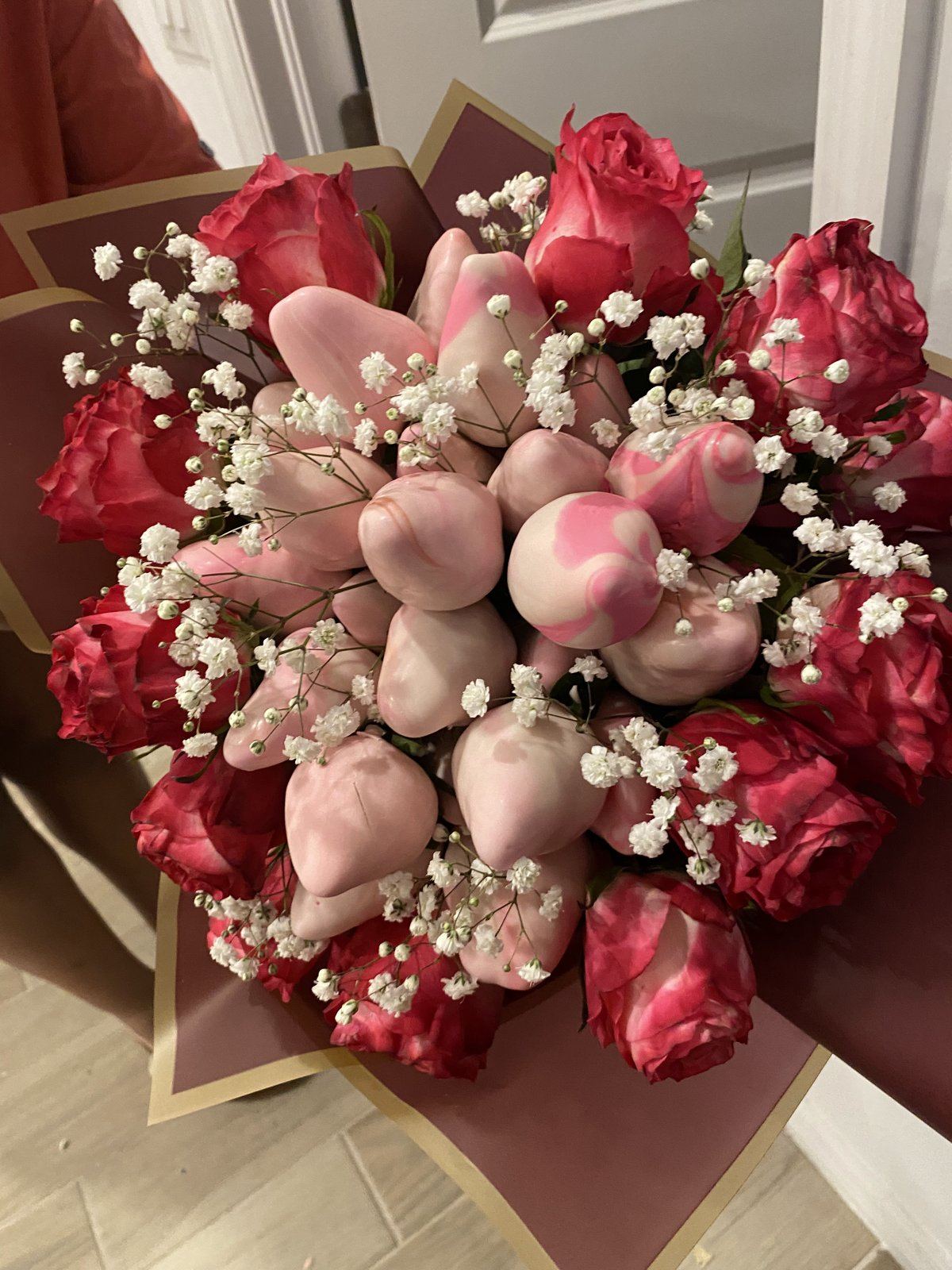 Bouquet of pink chocolate-covered strawberries surrounded by red roses and baby’s breath, wrapped in burgundy paper.