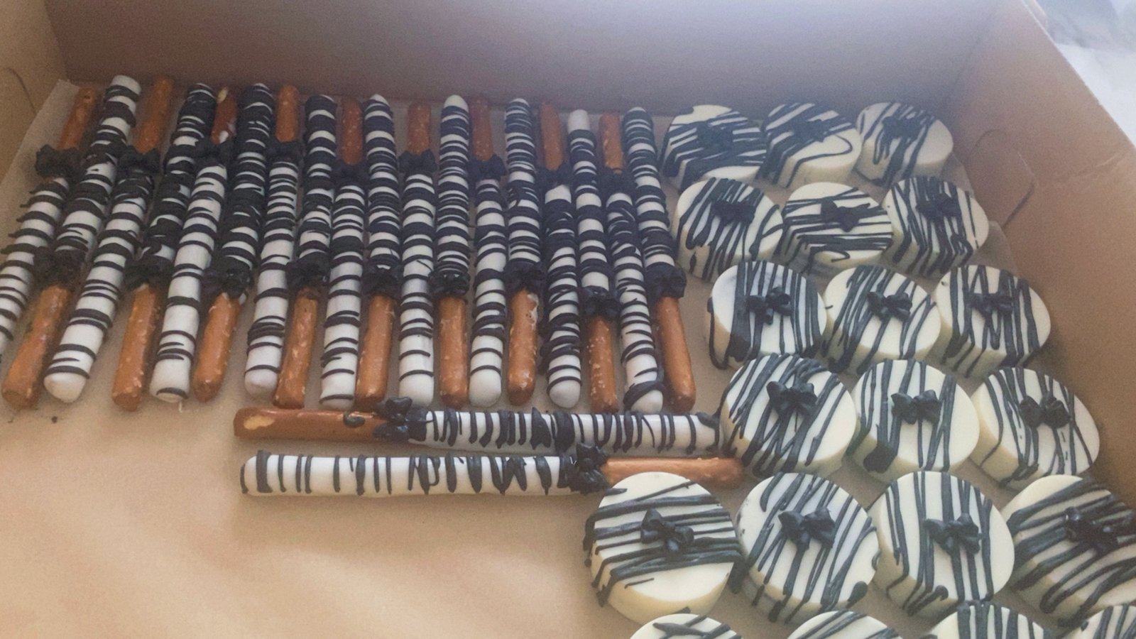 Box of white chocolate-dipped pretzel rods and cookies with black drizzle and small black bows.