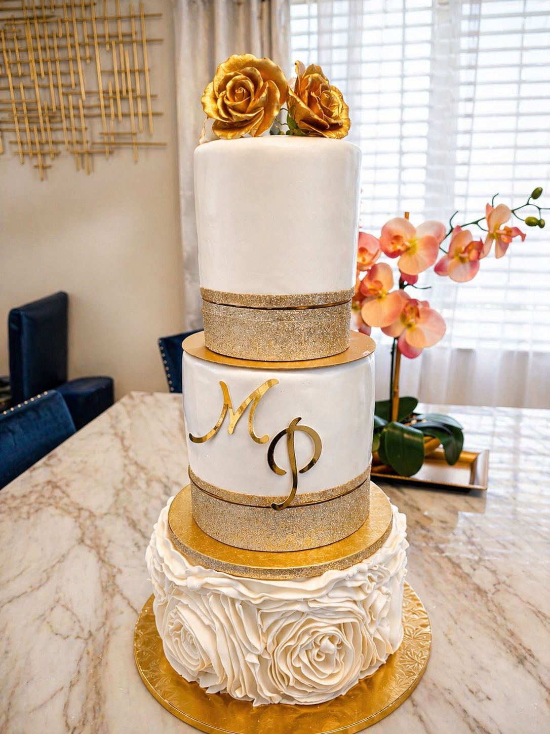 Three-tier white wedding cake with gold sugar roses on top, gold glitter bands, a gold MP monogram, and a ruffled white rose bottom tier.