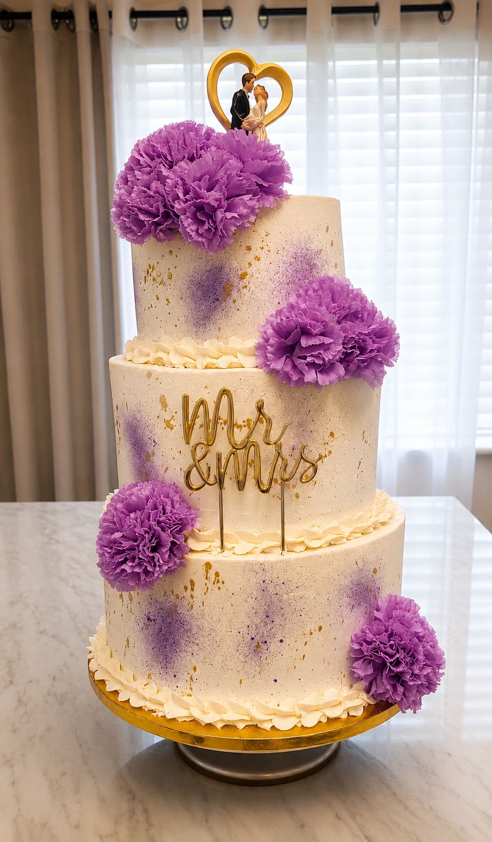 Three-tier white buttercream wedding cake speckled with lavender and gold, with purple carnations, a gold Mr & Mrs topper, and a couple figurine on top.