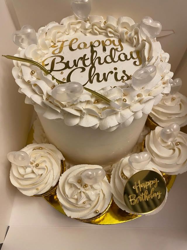 White birthday cake with gold Happy Birthday lettering, surrounded by matching white swirl cupcakes with gold pearls and clear heart toppers.
